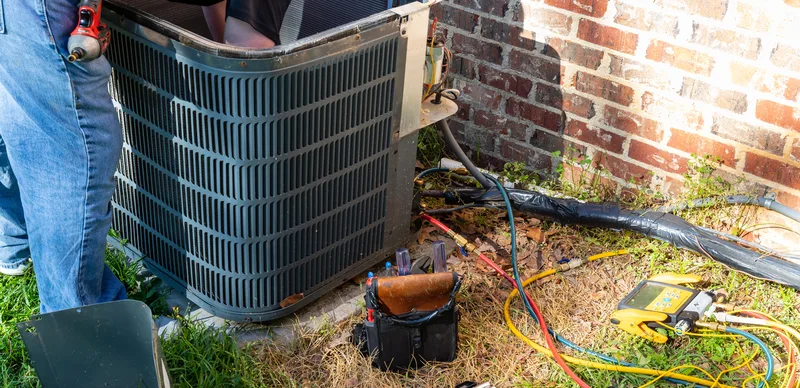 Technician's gauges connected to a residential central air conditioning condenser beside a brick home